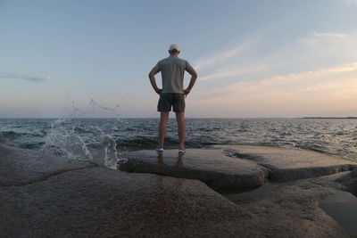 A man stands on a rocky seashore and looks to the horizon. back view