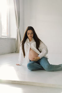 Cute young pregnant girl sitting on the white studio in warm light