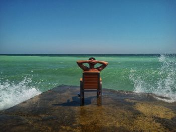 Rear view of mid adult man with hands behind head sitting on chair at beach against clear blue sky