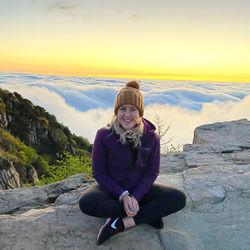 Portrait of young woman sitting on snowy field against sky during sunset