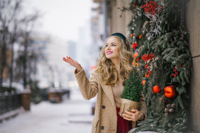 A pretty blonde holds a potted christmas tree in her hands, which she bought in a store, and walks