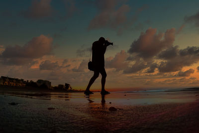 Rear view of man standing at beach against sky during sunset