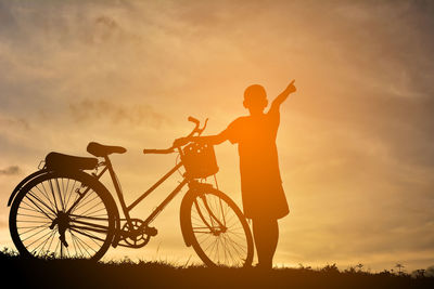 Silhouette boy with bicycle pointing towards sky while standing on field during sunset