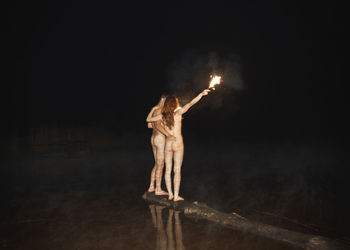 Rear view of naked couple holding flaming torch while standing on driftwood at lakeshore