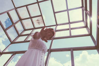 Low angle view of hand holding glass window against sky