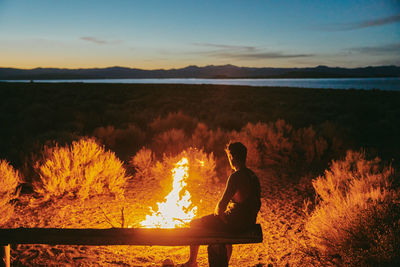 Reflection of man sitting on land against sky during sunset