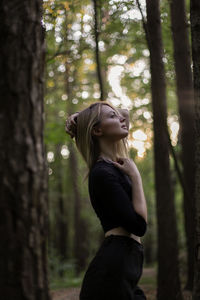 Side view of woman standing by tree trunk in forest