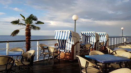 Empty chairs and tables at beach against sky