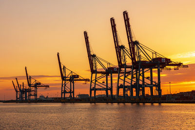 Cranes at commercial dock against sky during sunset