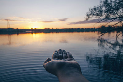 Cropped hand by lake against sky during sunset