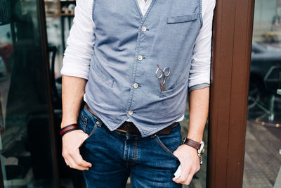 Cropped unrecognizable adult bearded male beauty master with hands in pockets at door of barbershop