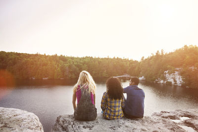 Rear view of friends sitting on rock by lake against clear sky