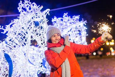 Portrait of smiling young woman with arms raised in city at night