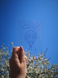 Cropped image of hand holding glass with drawing against tree and clear blue sky
