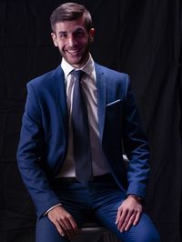 Portrait of smiling young man in suit sitting against black background