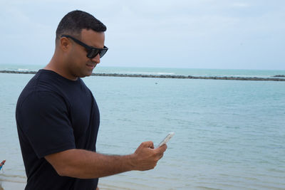 Man wearing sunglasses using mobile phone by sea against sky