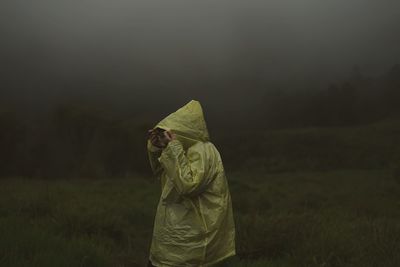 A woman standing and wearing yellow plastic jacket on field against misty cloud during rainy season