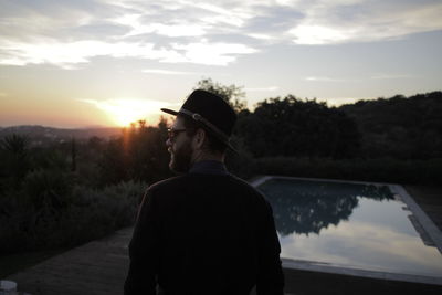 Rear view of man standing by pond against sky during sunset