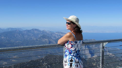 Woman standing by railing on mountain against blue sky