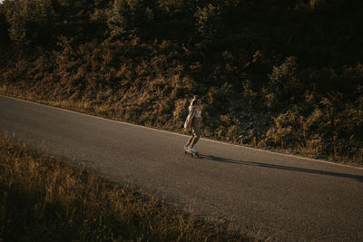 From above sporty female in trendy wear riding cruiser board along empty asphalt road in summer countryside on sunny day