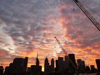 Silhouette cranes and buildings against sky during sunset