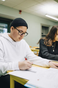 Teenage boy writing on book while studying at desk in classroom