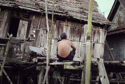 Rear view of shirtless man sitting on boardwalk by hut