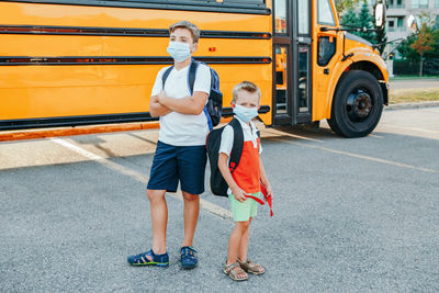 Brothers students in face masks standing by yellow school bus outdoor. 