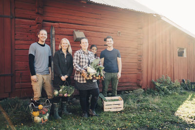 Full length of smiling farmers with organic vegetables standing against barn