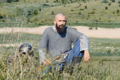 Viking. bald bearded man in metal chain mail over linen shirt sits on hill, holding ax. helmet lies