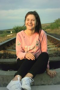 Portrait of smiling woman sitting on railroad track against sky