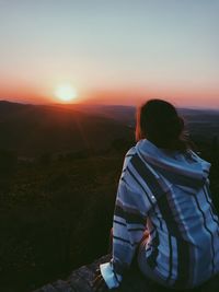 Rear view of woman sitting on cliff against sky during sunset