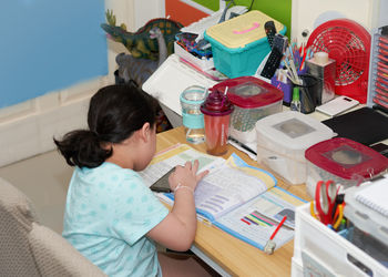 Elementary black haired girl doing her homework on the desk in her room at home high angle shot