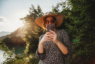 Woman with hat and sunglasses using mobile phone in sunset on the lake side