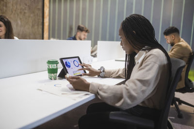 Side view of concentrated african american female employee using tablet while sitting at table and working in coworking space with multiracial colleagues
