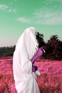 Man with covered blanket smoking cigarette on grassy field against sky