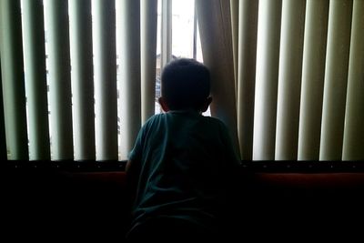 Rear view of boy looking through blinds window at home