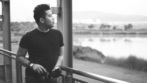 Young man looking away while standing by railing against sky