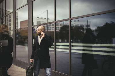 Smiling businessman talking on mobile phone while walking by city building