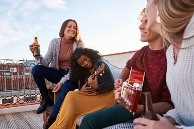 Cheerful young multiracial friends in casual wear playing acoustic guitar and ukulele while resting together on comfy sofa on building rooftop