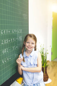 Portrait of smiling girl holding chalk while standing by blackboard in school