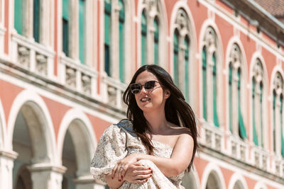 Portrait of beautiful young woman with long dark hair, sitting in square with beautiful architecture