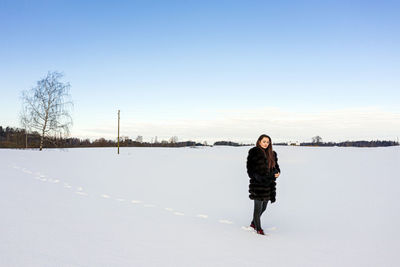 A cheerful woman in a warm fur coat walks across the open field leaving footprint in the snow