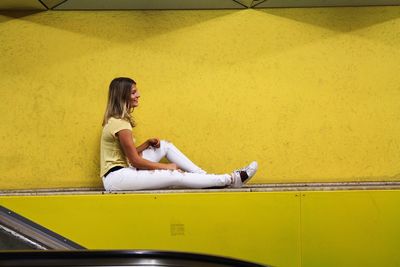 Young woman sitting on seat by yellow wall