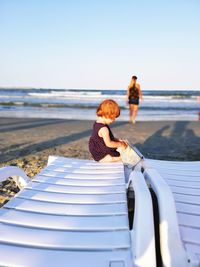 Girl sitting on lounge chair with mother in background at beach