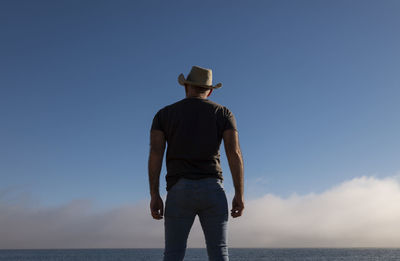 Rear view of adult man in cowboy hat and jeans against blue sky