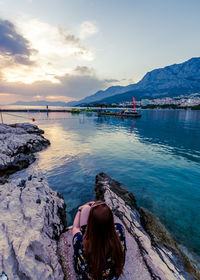 Rear view of mid adult woman looking at sea while sitting on rock against sky during sunset