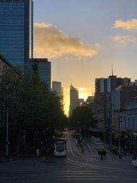 Cars on city street by buildings against sky during sunset