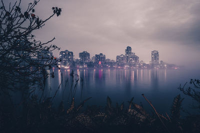 Illuminated city by river against sky at dusk