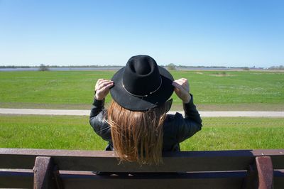 Rear view of woman wearing hat sitting on bench at field against clear blue sky during sunny day